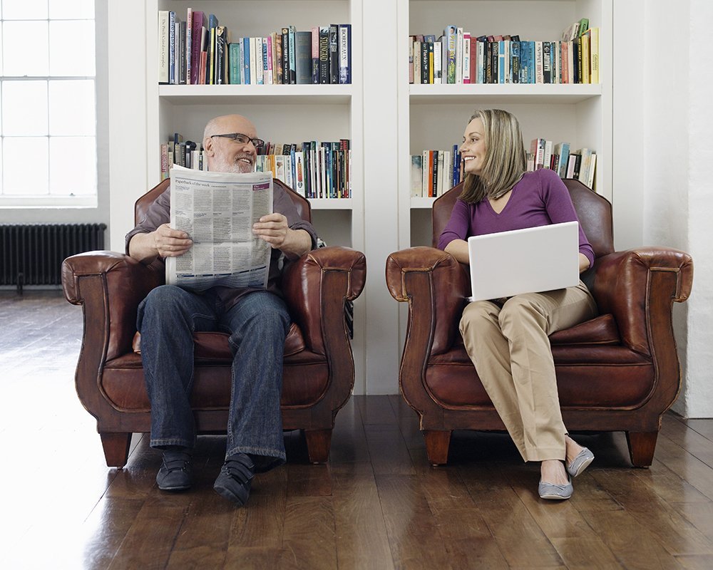 Full length of a middle aged couple sitting in armchairs with newspaper and laptop at home